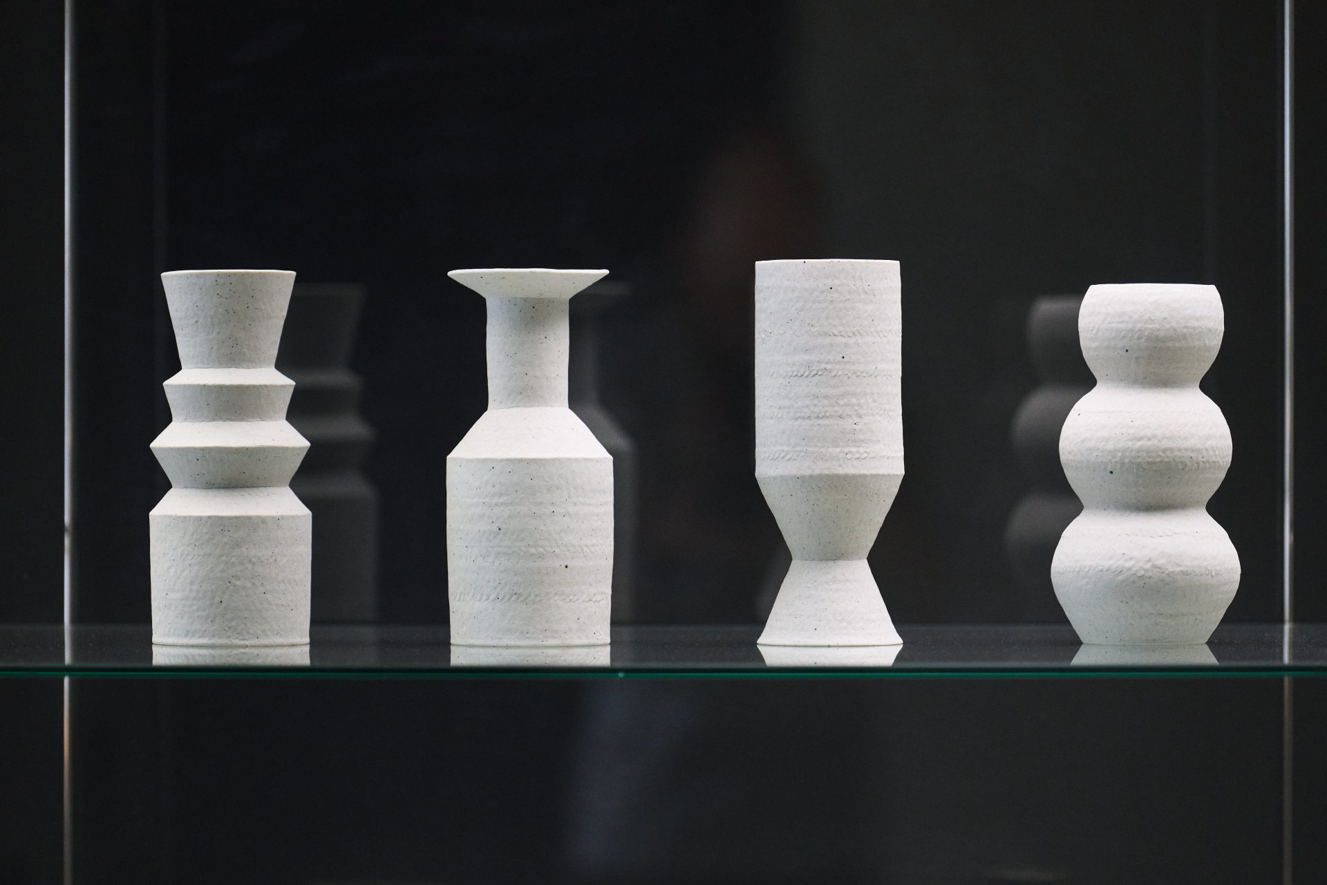 Vier moderne Vasen in verschiedenen Formen und Höhen aus hellem Material auf einem Glasregal.