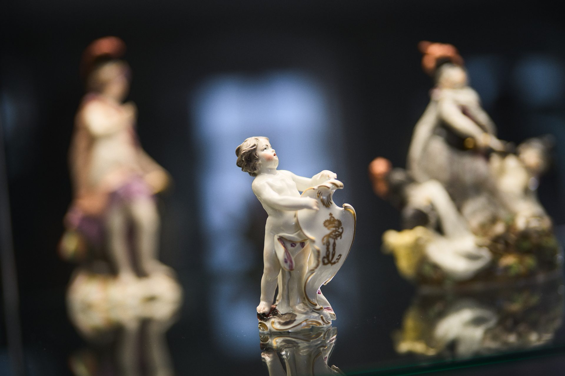Porzellanfigur eines nackten Kindes mit Schild, im Hintergrund weitere Figuren.