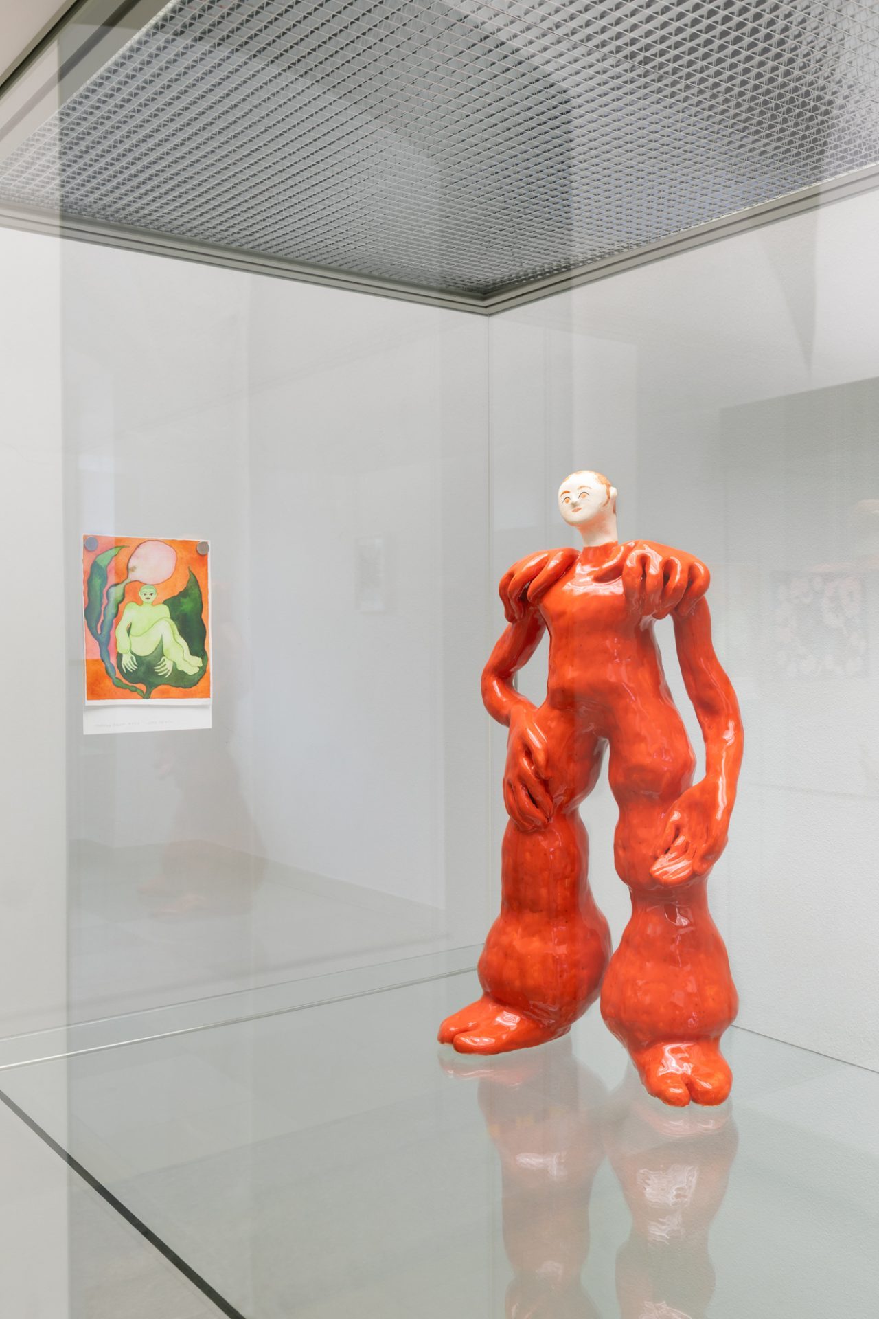Skulptur in leuchtendem Orange mit übergroßen Armen und Beinen hinter Glas, daneben ein buntes Bild.