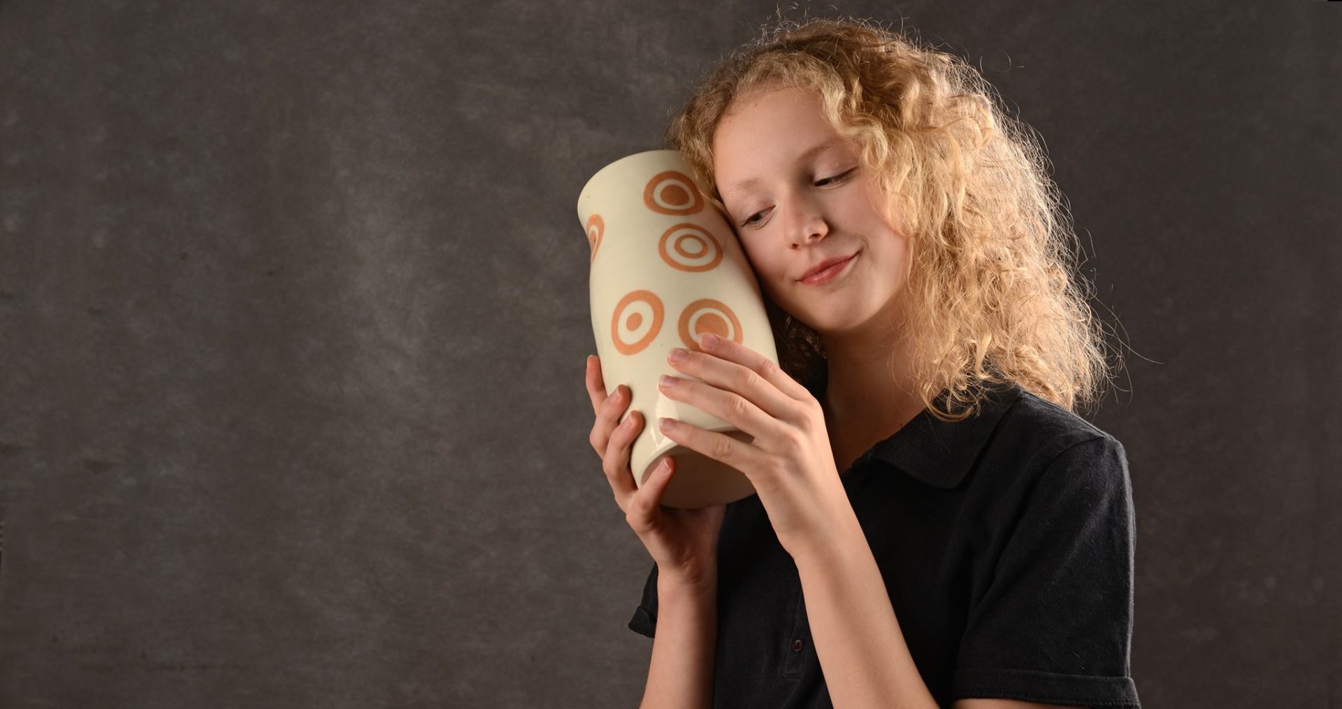 Mädchen mit lockigem Haar, das liebevoll eine Vase mit kreisförmigen Mustern vor dunklem Hintergrund hält.