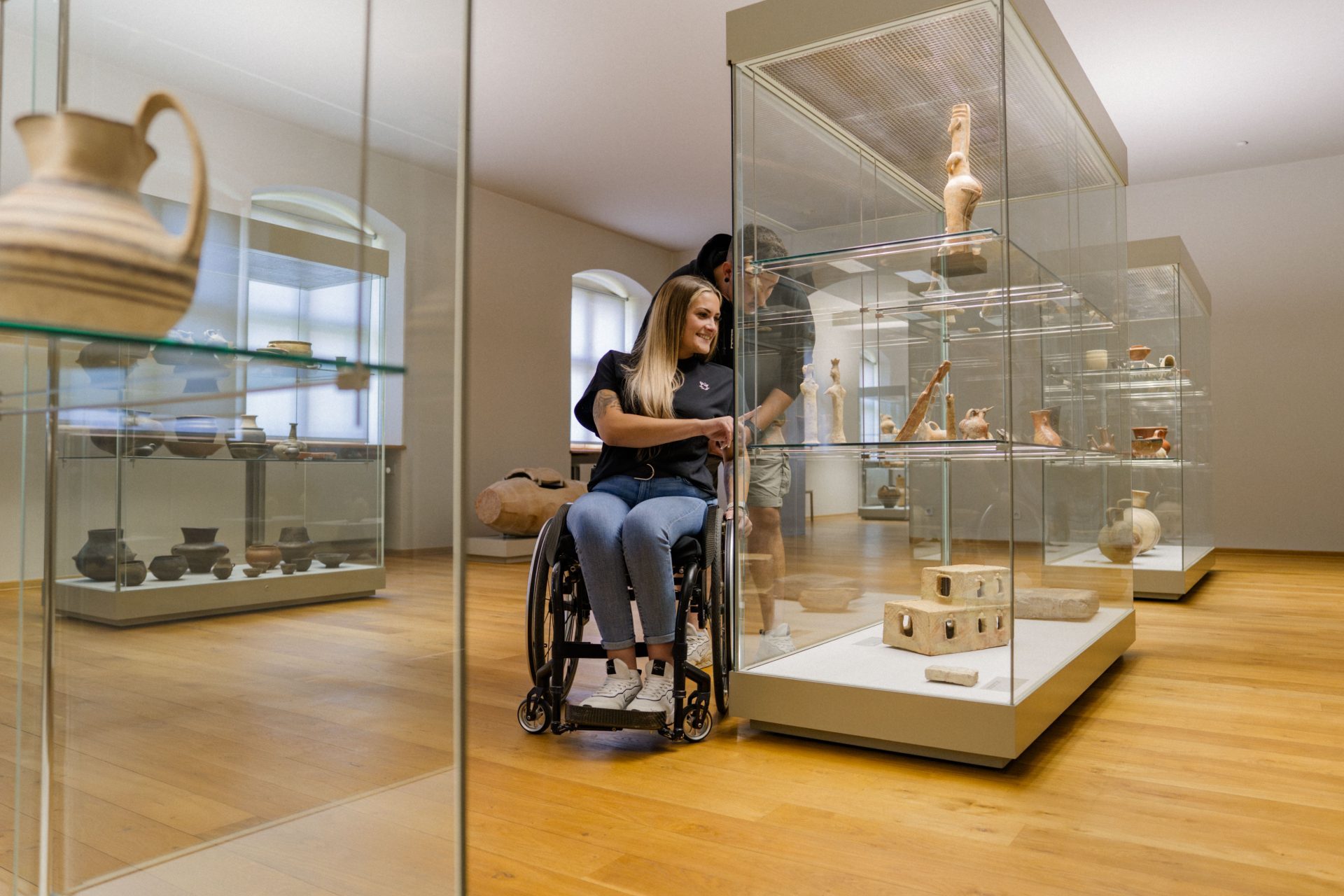 A young woman in a wheelchair examines ancient artifacts in a museum with a companion.