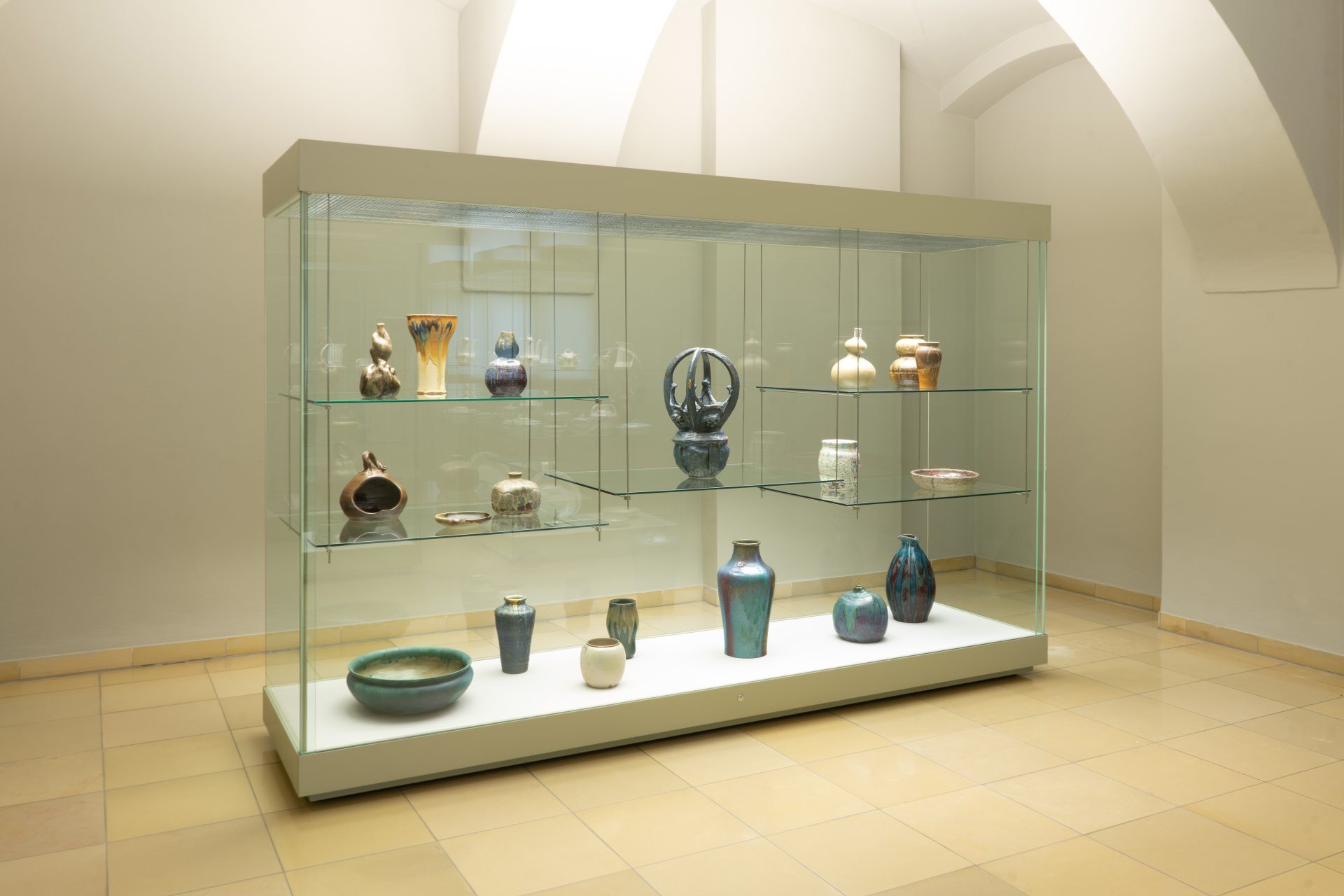 Glasvitrine mit bunten Kunstwerken und Vasen in einem lichtdurchfluteten Ausstellungsraum.