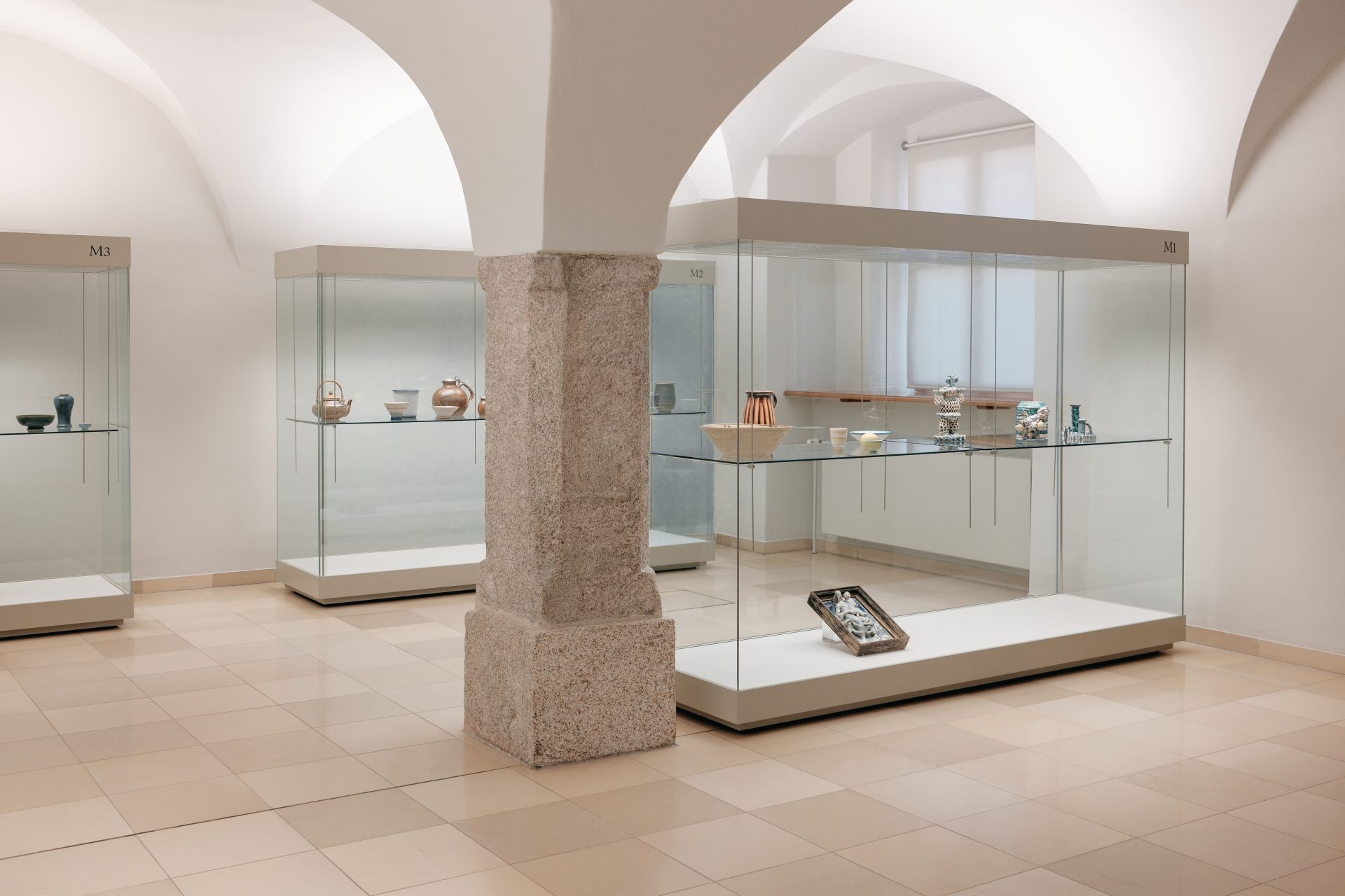 Moderne Ausstellung mit Vitrinen, die keramische Objekte in einem hellen Raum unter einem architektonischen Bogen präsentieren.