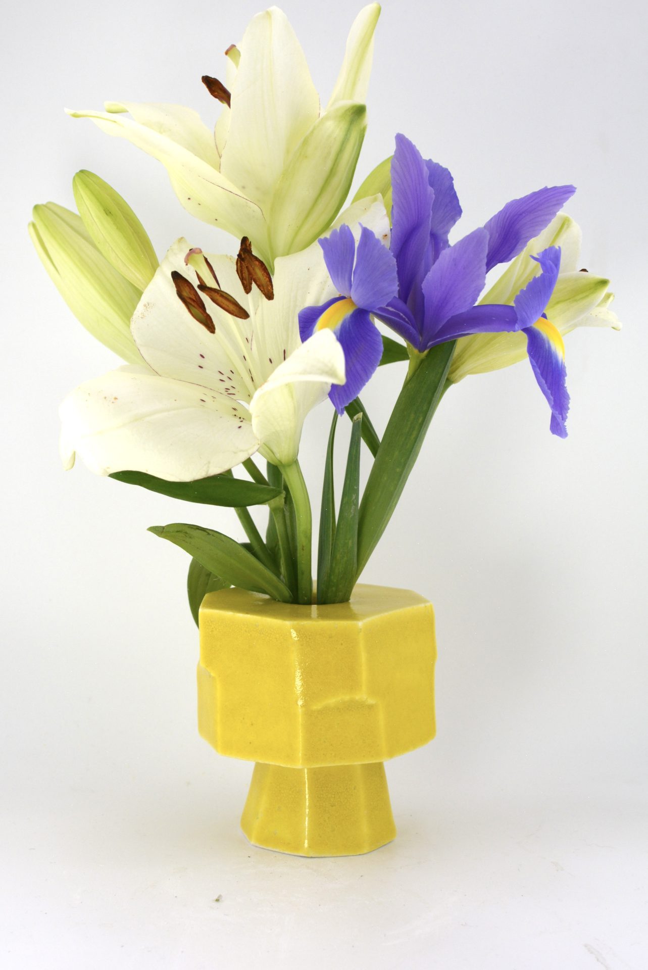 Buntes Blumenarrangement mit weißen Lilien und lila Iris in einer gelben, geometrischen Vase.